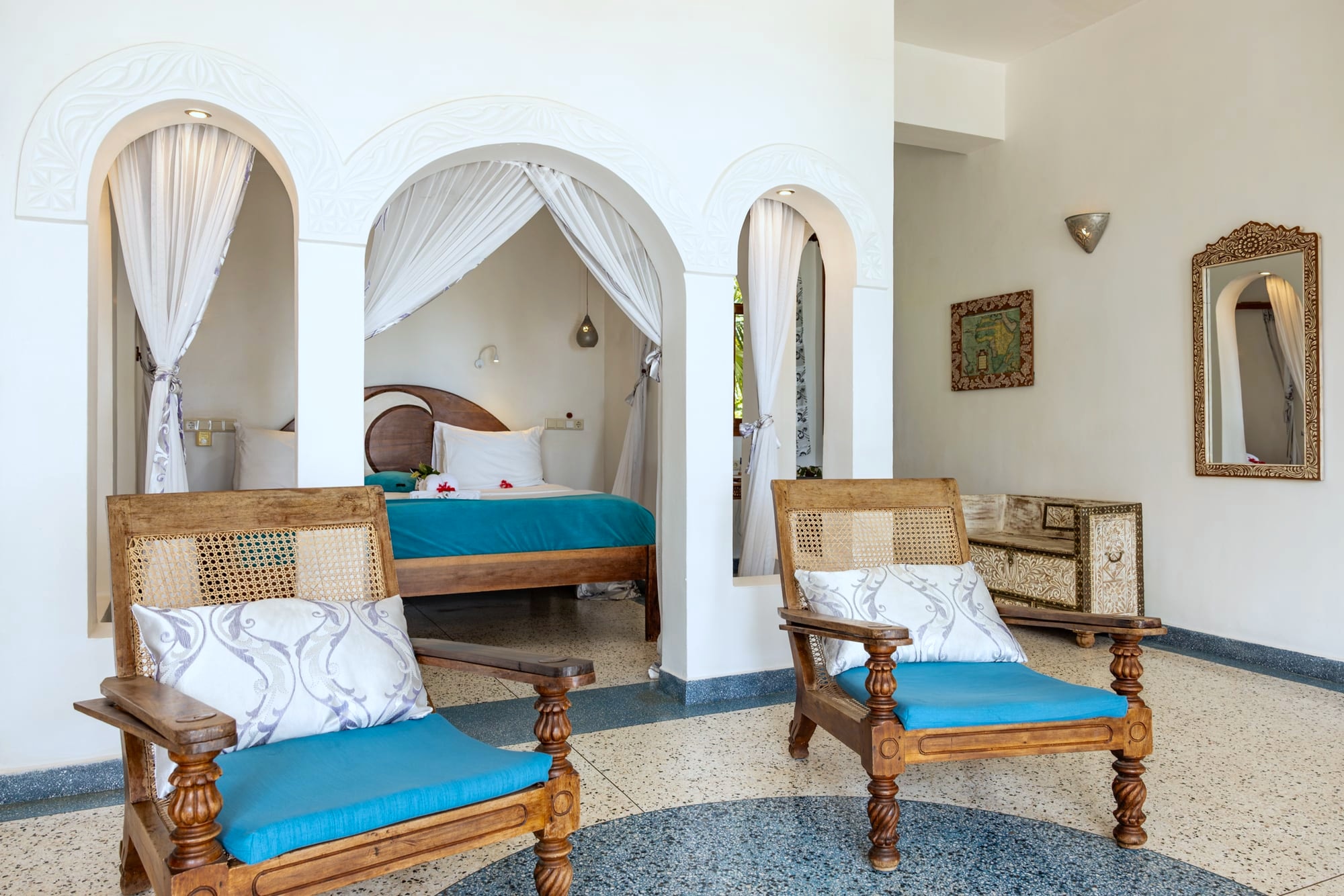 La suite Mwezi de Villa Kidosho: sillones de plantador tallados con cojines turquesa sobre suelo de terrazo, con alcobas en arco al fondo.