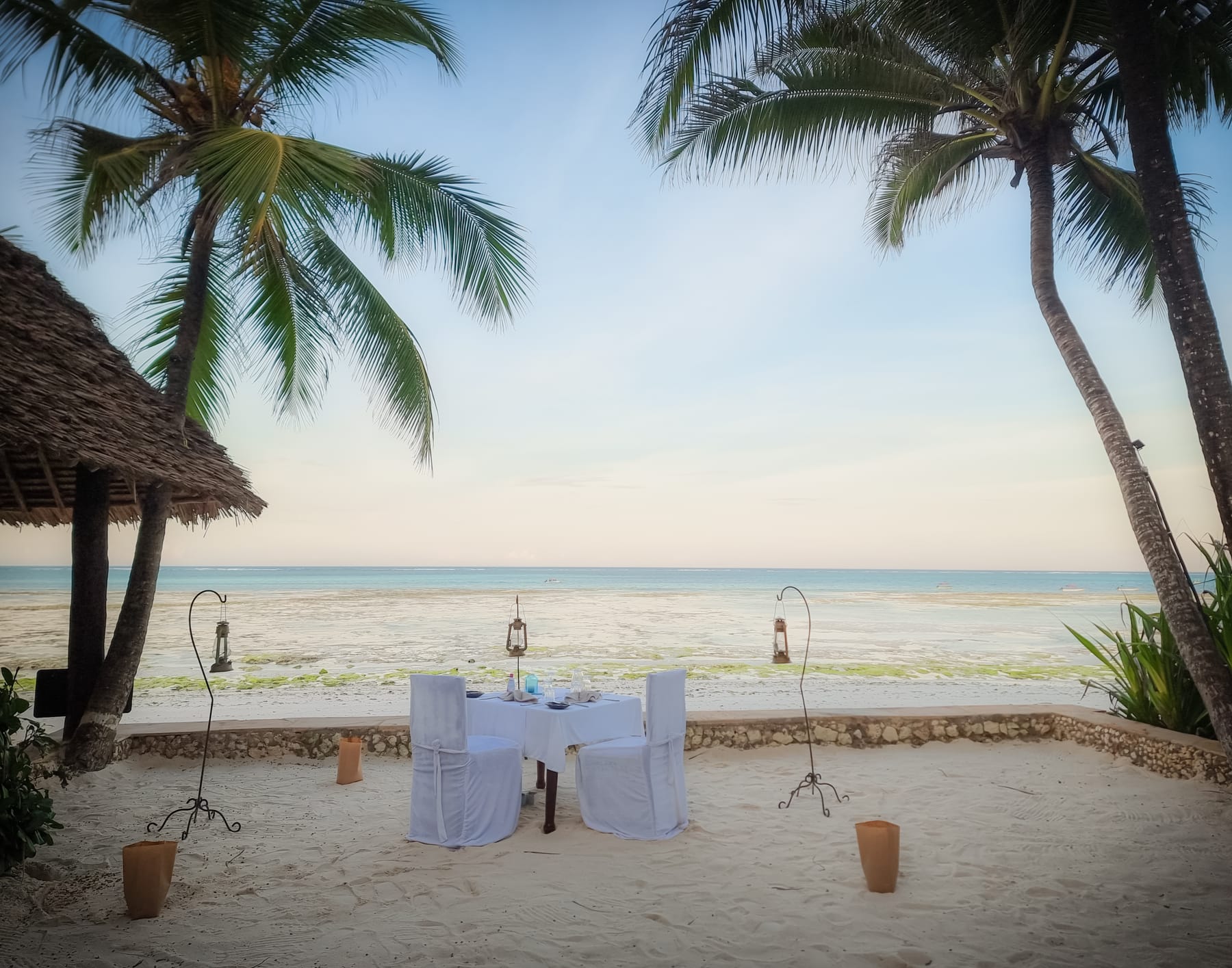 A table for two set on the sand at dusk, between palms, with lanterns