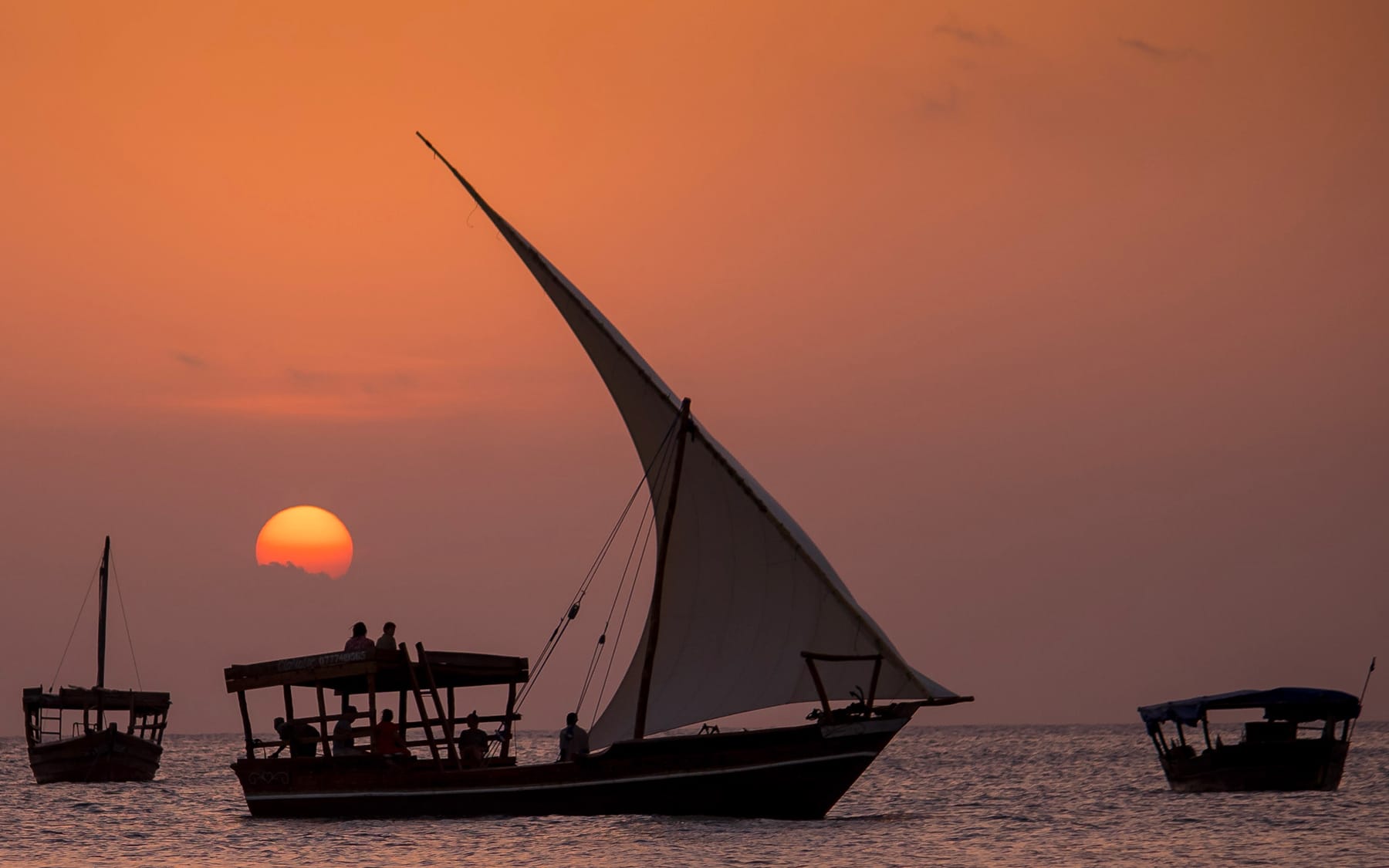Dhow boats sailing at sunset off Zanzibar, people silhouetted on the shore as the evening call to prayer nears