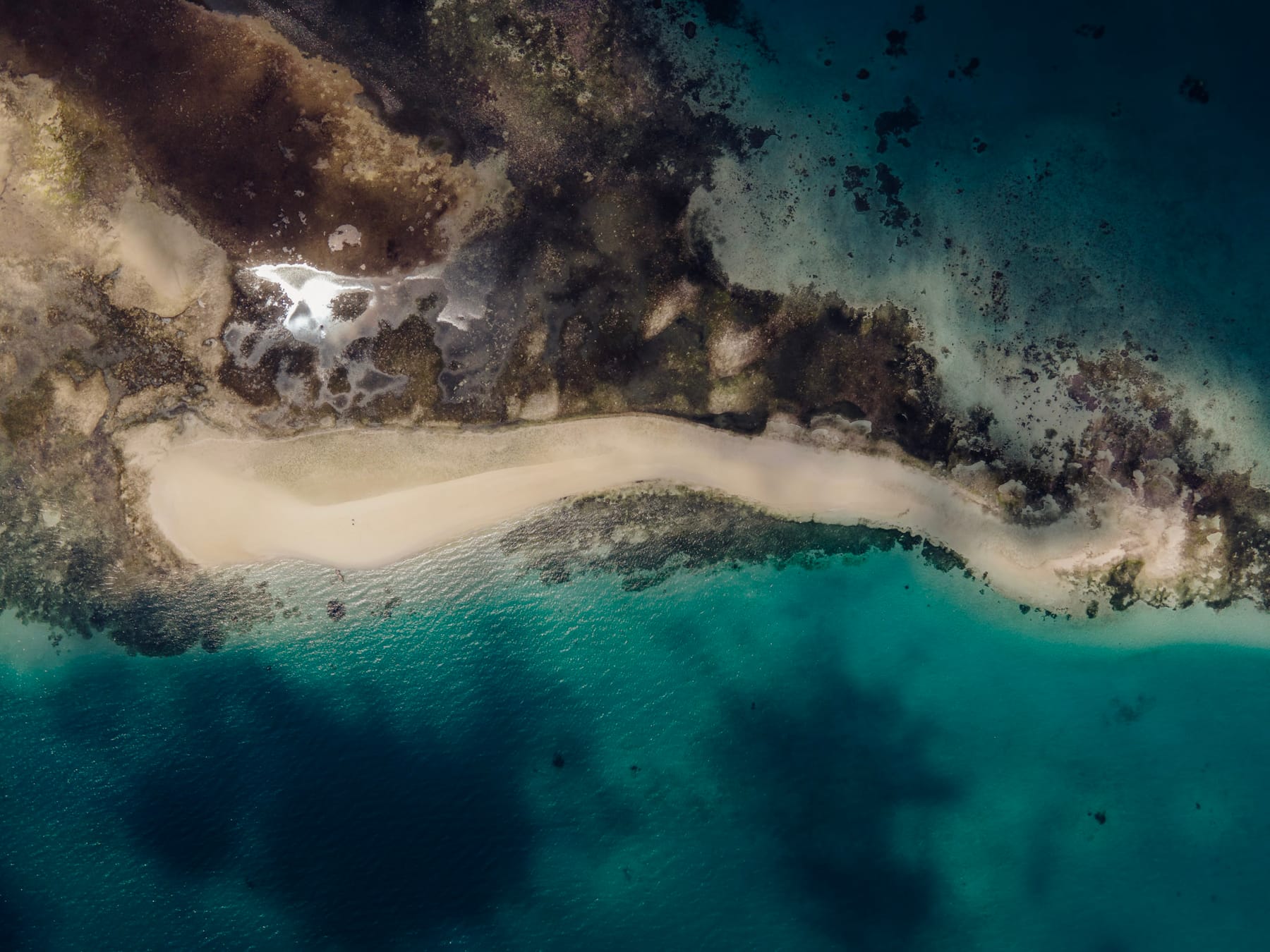 A pale sand spit running from a rocky, reef-lined shore into turquoise water, seen from above