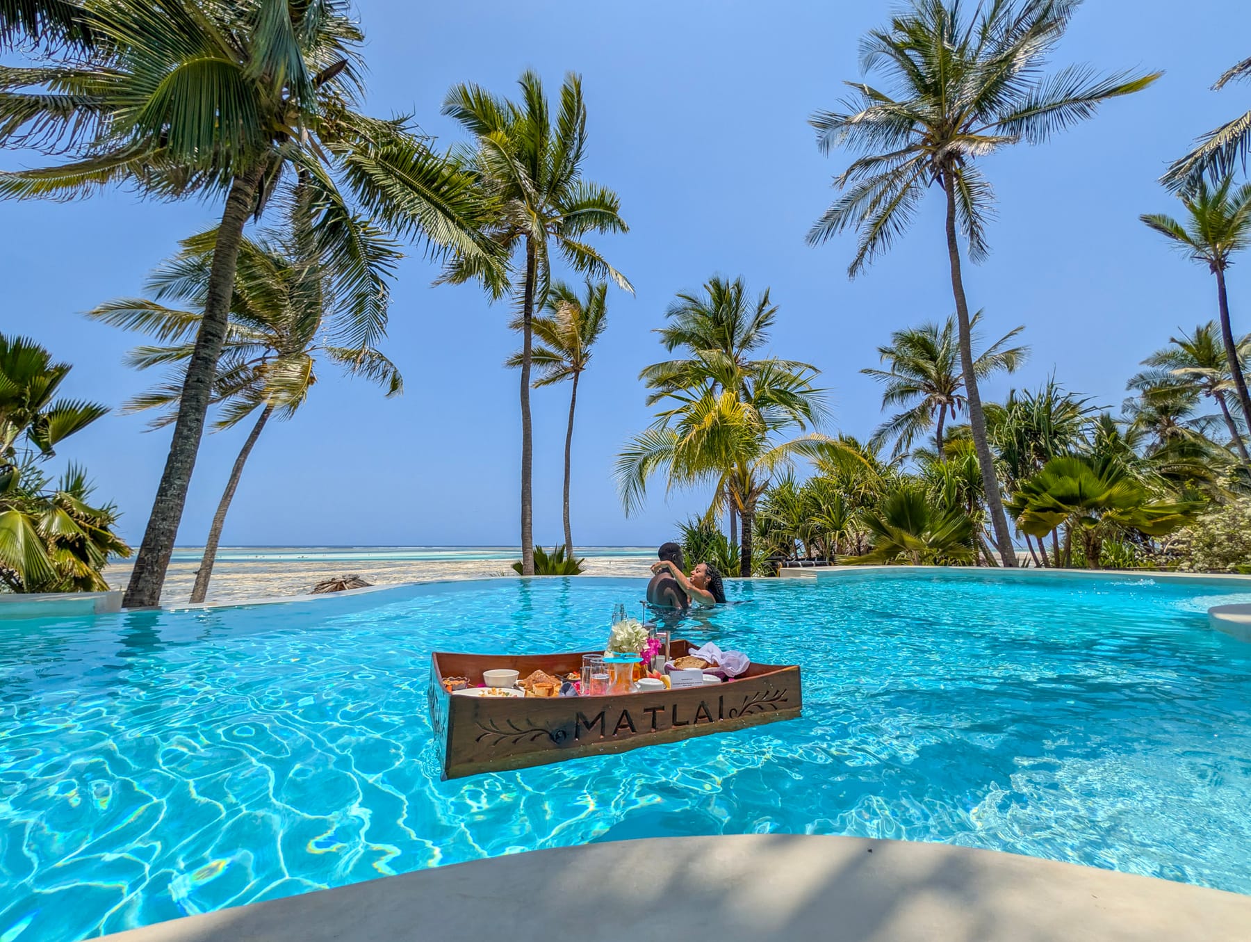 A wooden Matlai tray floating on the infinity pool, laid with fruit and pastries, a couple in the water behind it