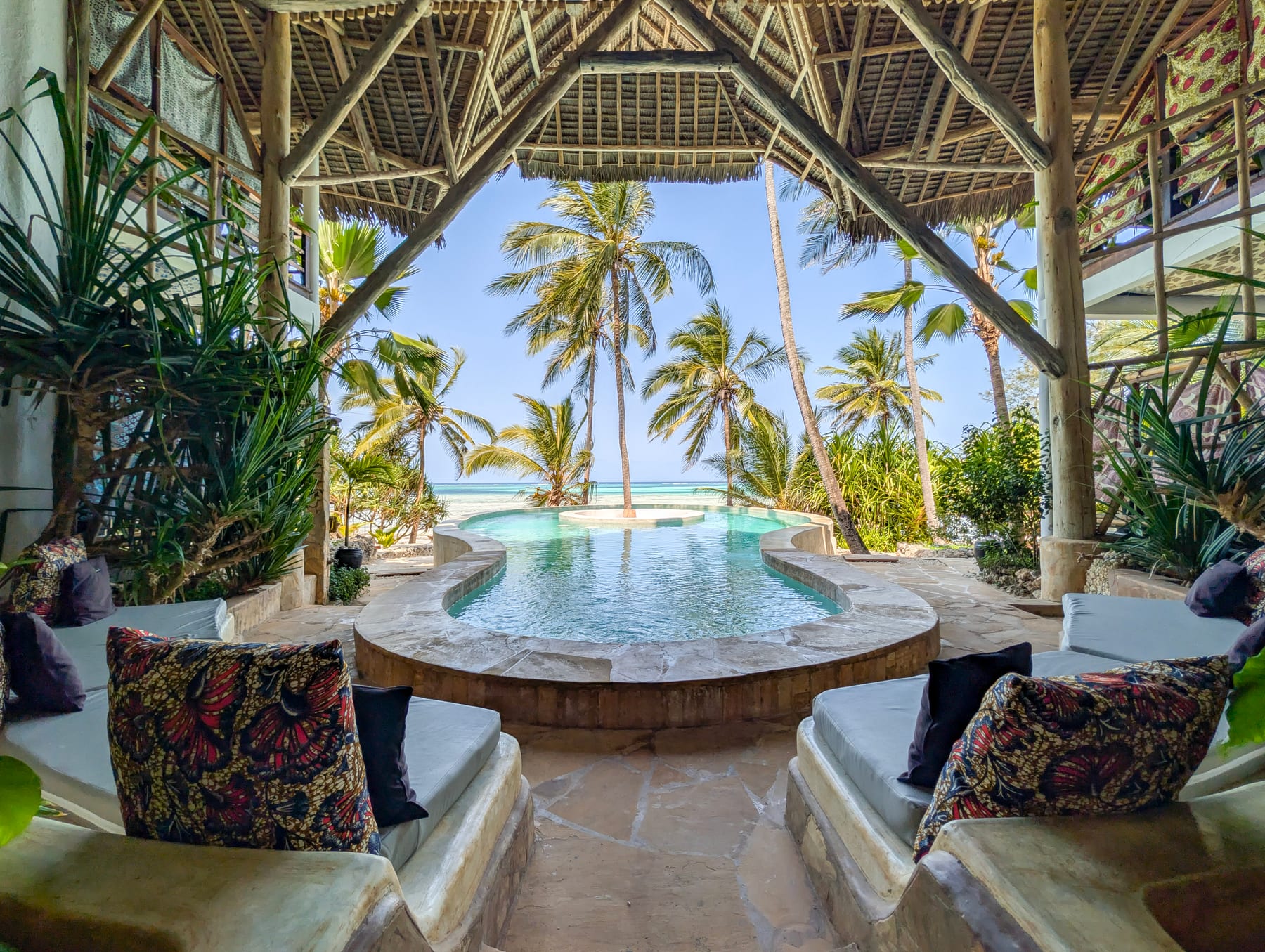 Cushioned seating under a thatched roof at Asili House, with the pool and palm trees beyond