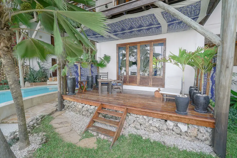 The covered wooden terrace of Room Maji with a dining table and chairs under a blue patterned ceiling, potted palms and steps down to the garden