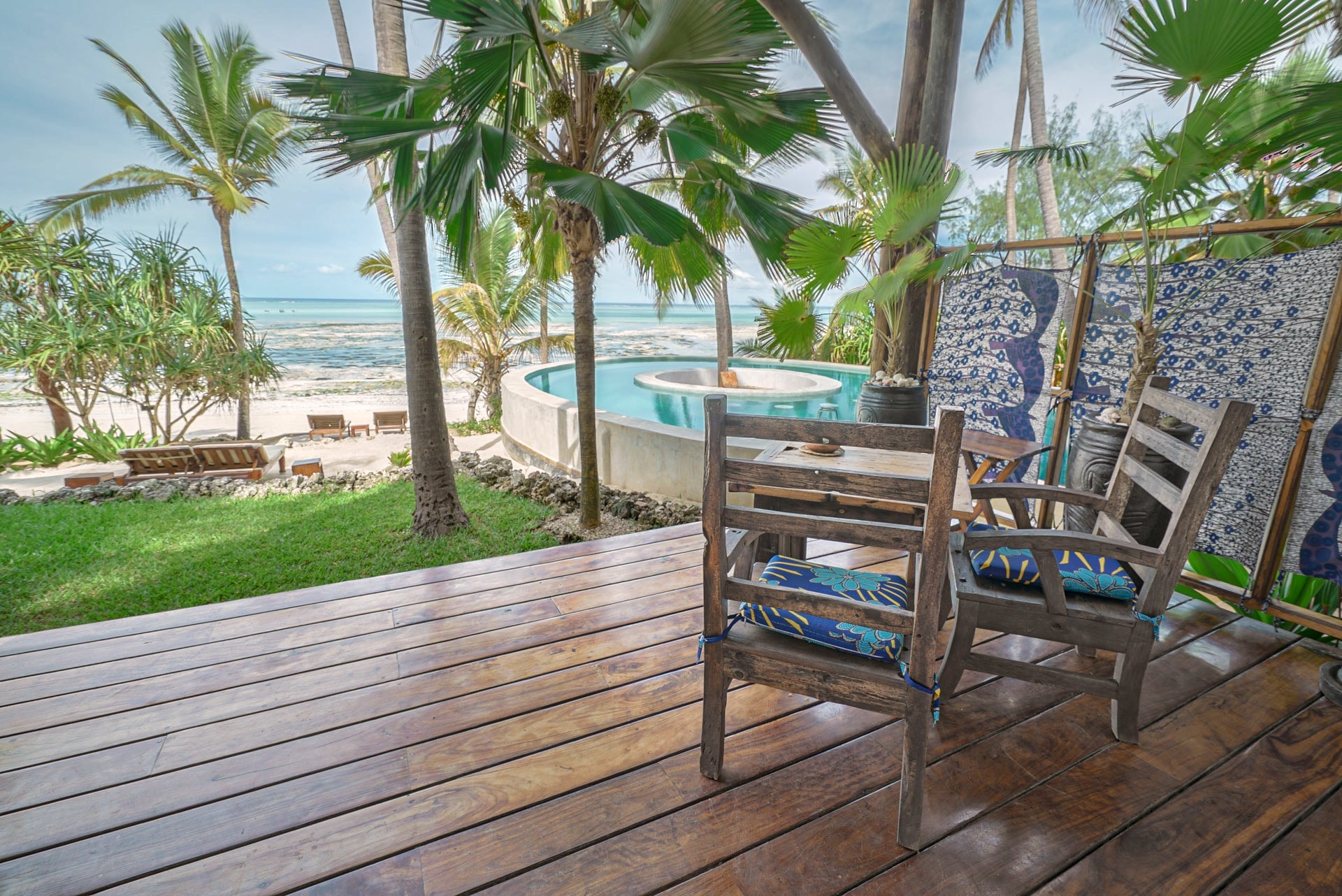 Deux chaises en bois aux coussins bleus sur la terrasse de la chambre Maji, avec des palmiers, la piscine et l’océan au loin