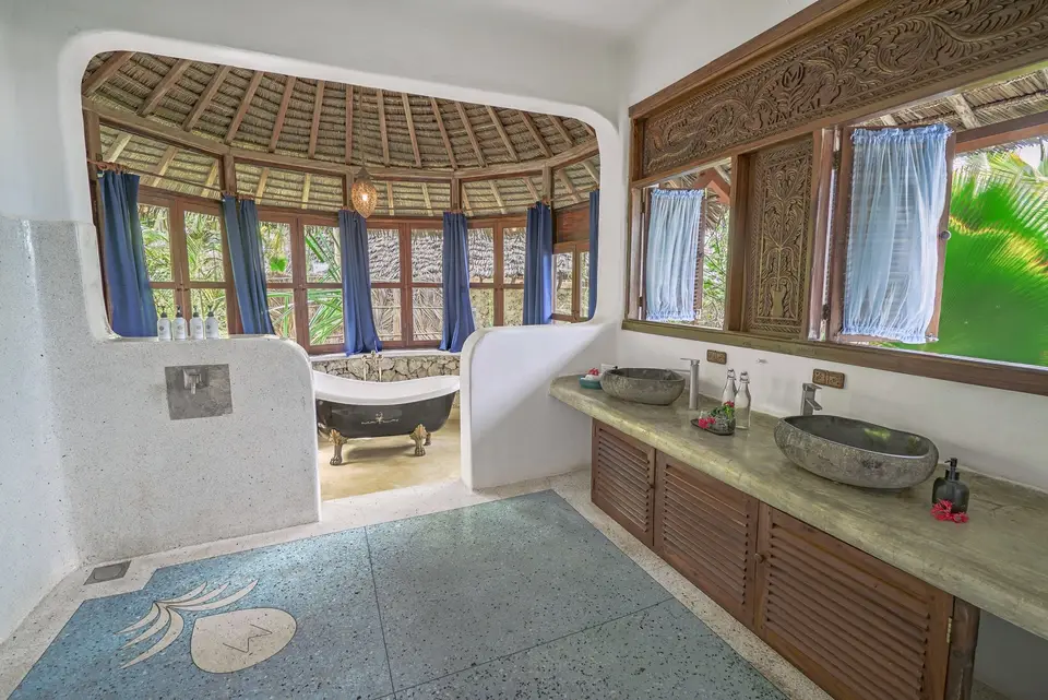 The round, thatch-roofed bathroom of Room Maji at Asili House, Boutique Hotel Matlai in Michamvi Pingwe: a long wooden vanity with two stone basins, a freestanding bathtub in the centre and blue curtains at the windows.