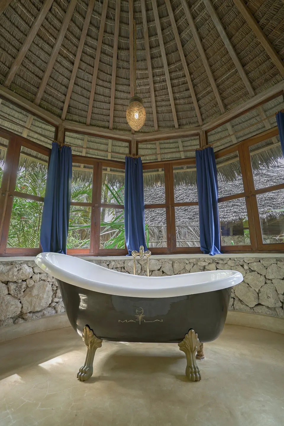 A freestanding clawfoot bathtub under a thatched roof in the bathroom of Room Maji, with blue curtains at the windows