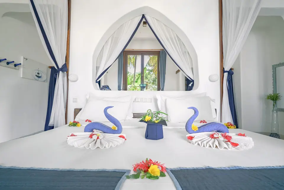 A white bed with blue accents under a mosquito-net canopy in Room Maji, with towel swans and fresh flowers