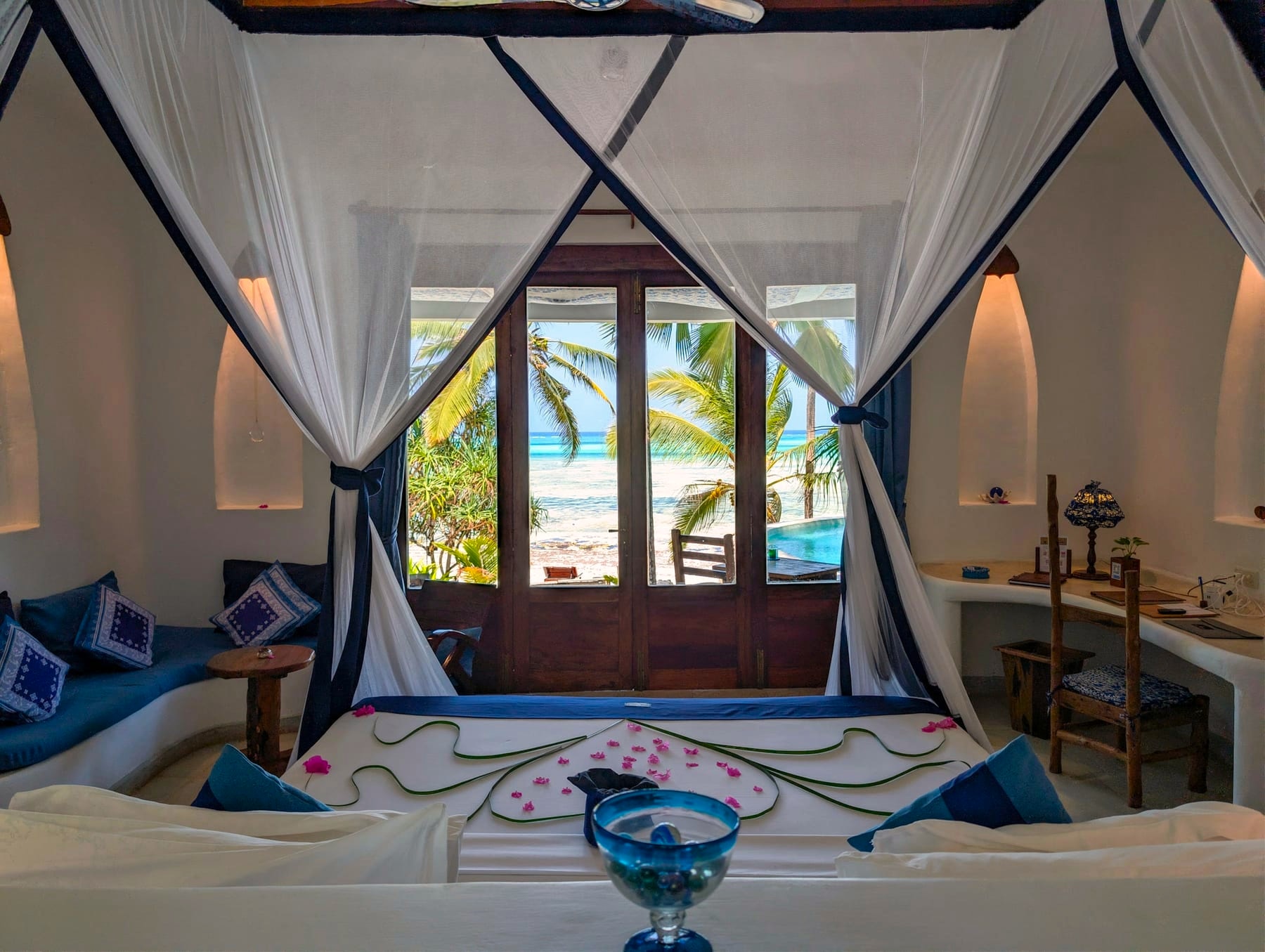 Un letto a baldacchino con zanzariera e cuscini blu in una camera di Asili House, con le porte aperte su palme e mare