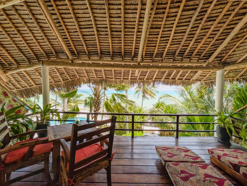 The thatched balcony of Room Moto at Asili House with kanga-print daybeds, a table and chairs, and palm trees and the ocean beyond