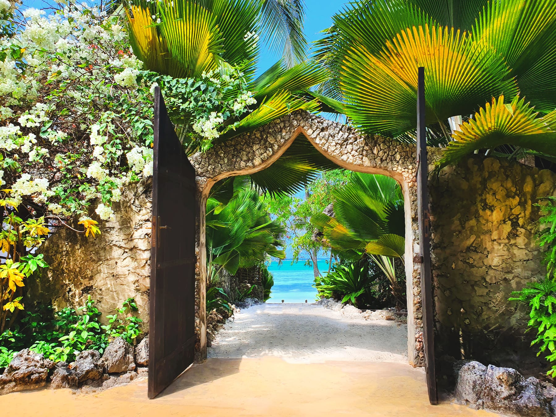 An open wooden gate in a stone wall, with a sandy path through palm fronds leading to the turquoise sea