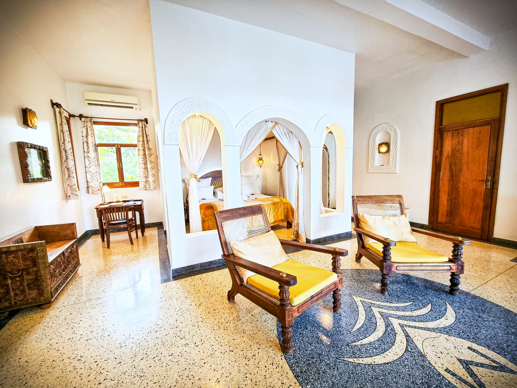 The Jua suite at Villa Kidosho — carved planter's chairs and the four-poster bed beyond arched openings, on a terrazzo floor
