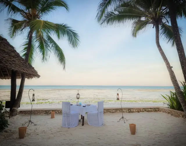 A table with a white cloth set on the sand under palm trees, with the ocean and torches behind it
