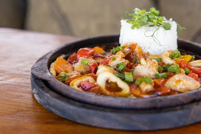 Close-up of a sizzling plate with prawns, squid and vegetables and a mound of white rice with herbs
