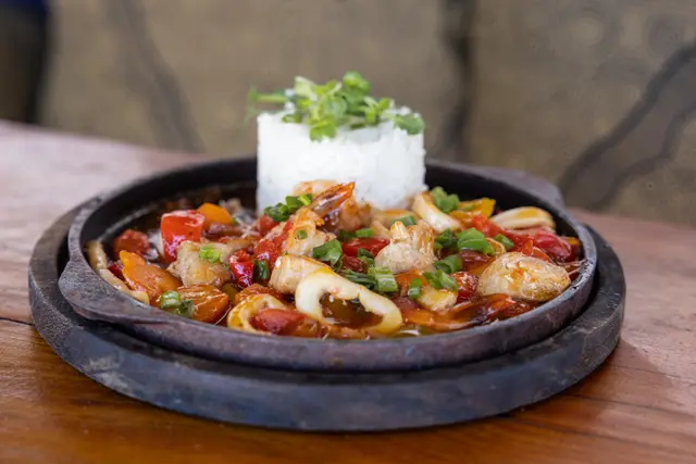 A sizzling plate of seafood and vegetables with a mould of rice on top, on a table outdoors