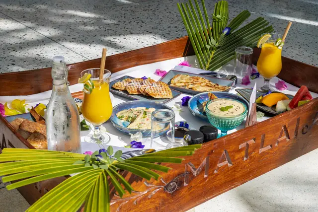 A wooden floating tray with juice, bread and small bowls, laid with a palm leaf, on the blue water of the pool