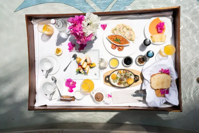 A breakfast tray seen from above with fruit, pastries, small dishes and pink flowers, floating on pale water