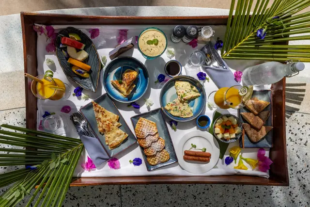 A breakfast tray seen from above, with bowls, pastries and flowers arranged between palm leaves