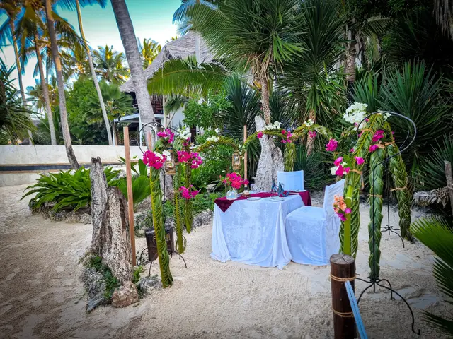 A table with a white cloth set at the end of a path lined with flowers and tall flowering arches, among palm trees