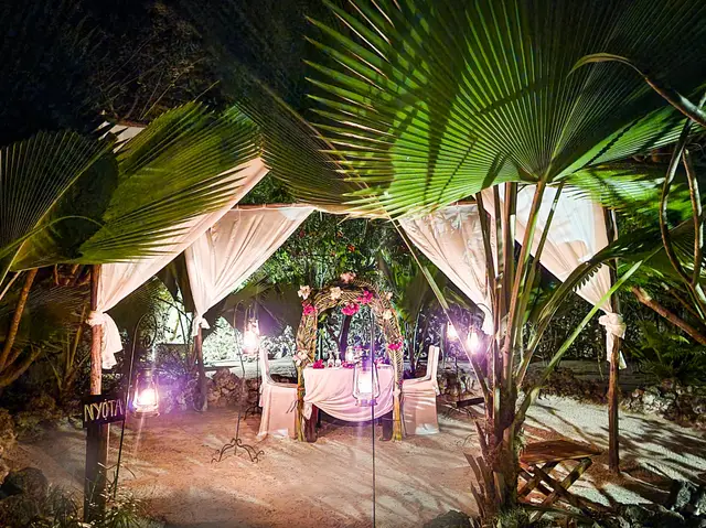 A table set for two under a draped canopy among palm leaves, lit by candles and soft purple lights at night