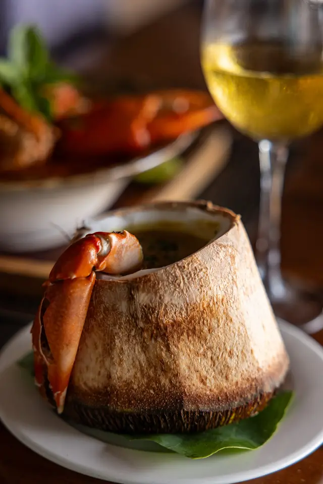 Soup served in a coconut with a crab claw resting on it, beside a glass of white wine