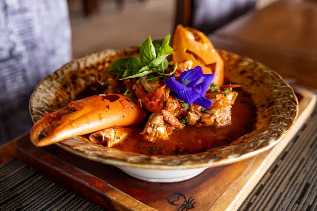 A crab dish with claws in a rich orange sauce on a wooden serving board, decorated with a purple flower