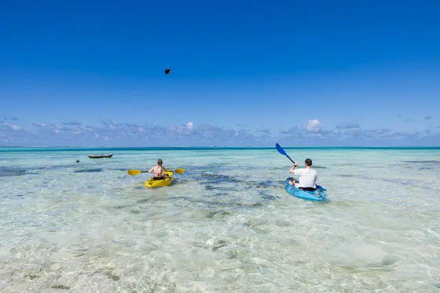 Two small figures in kayaks on very shallow clear water, with sand ripples and rocks showing below