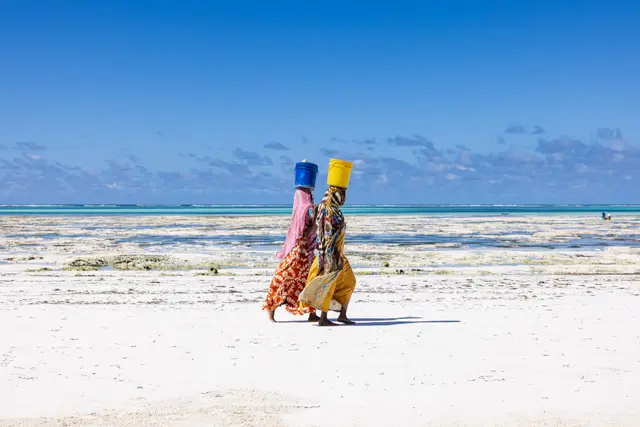 Two women walking along a white sand beach, seen from behind, carrying buckets on their heads