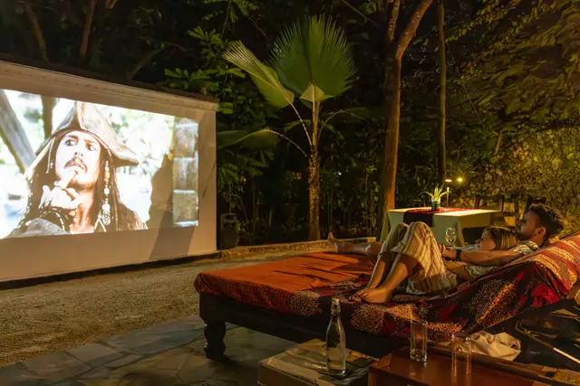 A film on a large outdoor screen in the garden at night, with a daybed in front