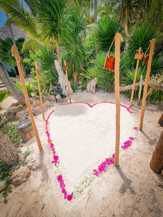 A heart outlined in pink flower petals on white sand between wooden poles, set up for a proposal