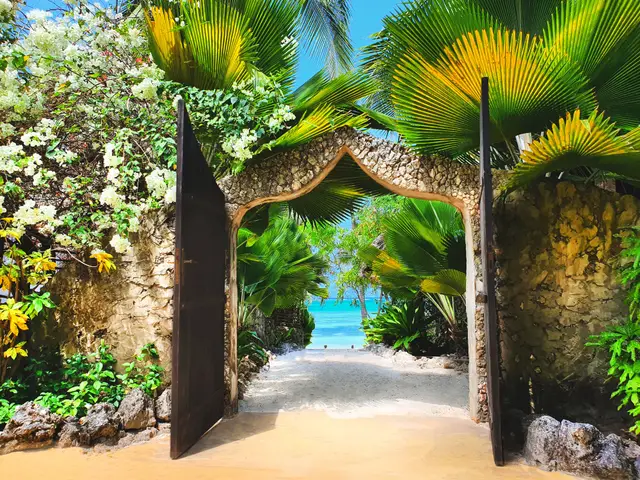 An open wooden garden gate in a stone wall, with a sandy path leading through tropical plants to the turquoise sea