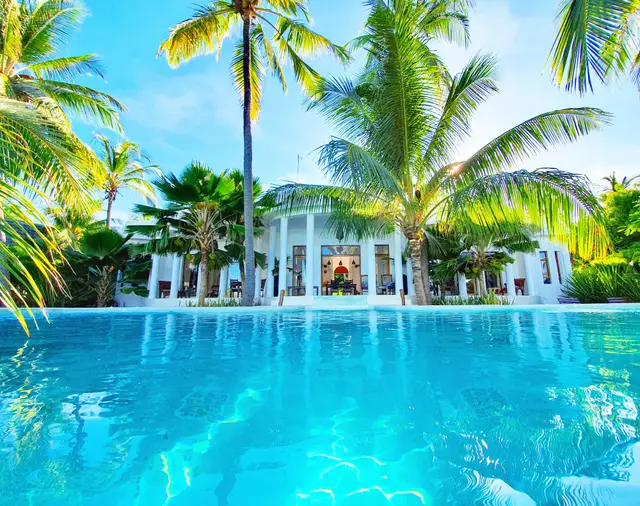 A turquoise pool with a white colonnaded villa and tall palm trees behind it in the afternoon sun