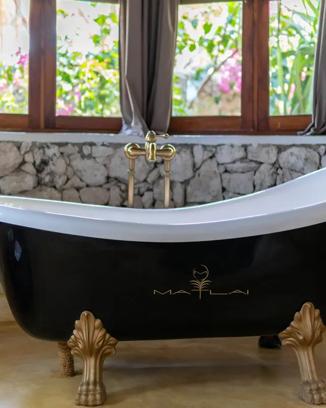 A black freestanding bathtub with gold-coloured claw feet in front of a stone wall and tall windows in the Dunia room