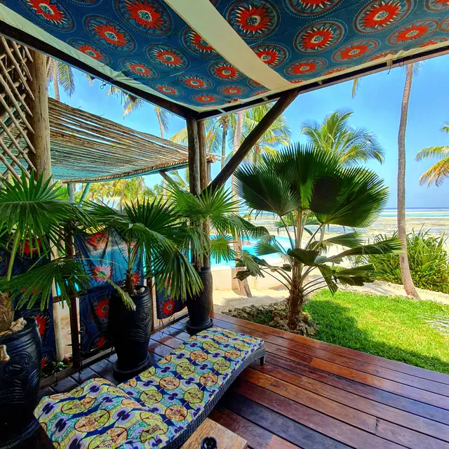 A covered wooden terrace with a daybed under a colourful patterned ceiling, with a view of the lawn and the ocean