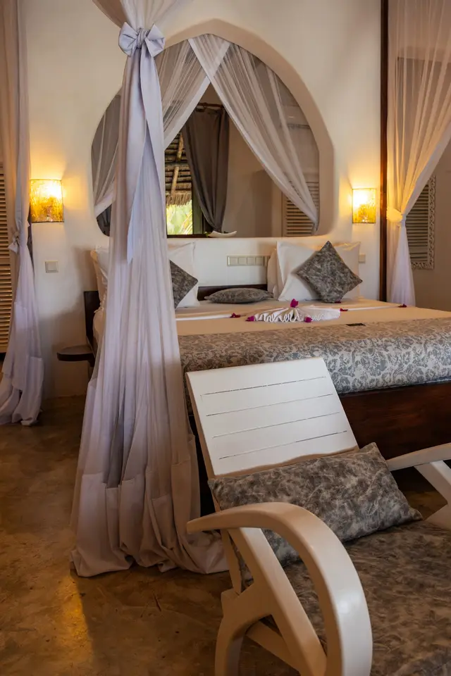 A bed under white mosquito nets with a wooden lounge chair in front, in the Hewa room