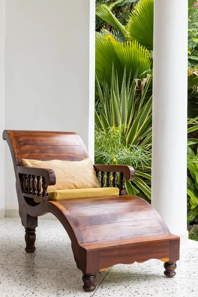 A carved wooden lounger on the balcony of the Jua suite between white columns, with garden leaves behind
