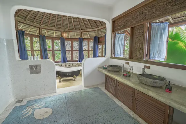 The round, thatch-roofed Maji bathroom: a long wooden vanity with two stone basins, a freestanding bathtub in the centre and blue curtains at the windows