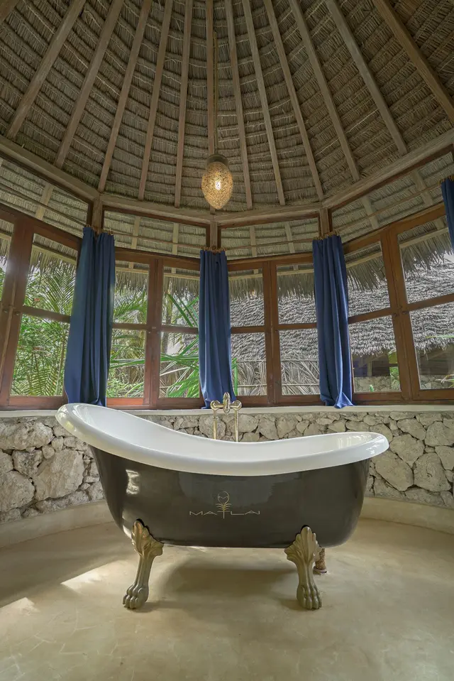 A freestanding clawfoot bathtub under a thatched roof in the Maji bathroom, with blue curtains at the windows