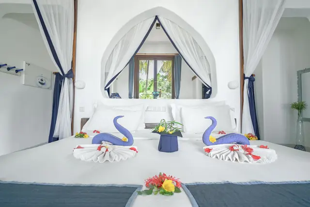 A white bed with blue accents under a mosquito-net canopy in the Maji room, with towel swans and fresh flowers