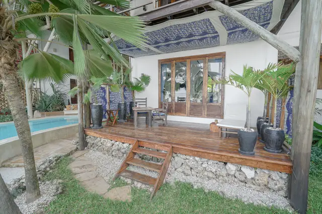 The covered wooden terrace of the Maji room with a dining table and chairs under a blue patterned ceiling, potted palms and steps down to the garden