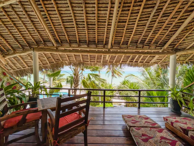 The Moto balcony under a palm-leaf roof with a table, chairs and kanga-print daybeds, and palm trees and the ocean beyond