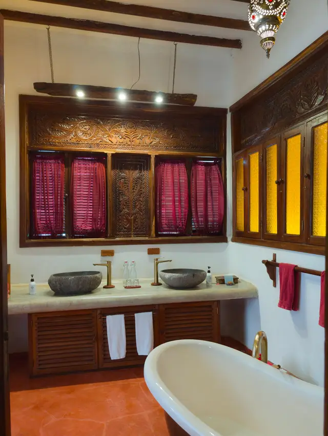 The Moto bathroom: a carved wooden panel with red curtains above a wooden vanity with two stone basins, amber glass windows and a white bathtub