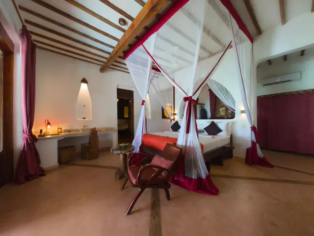 The bed under a mosquito net in the Moto room seen from the side, with red and white drapes, a wooden armchair with a red cushion and a lit lamp