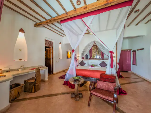 A wide view of the Moto bedroom: a bed under a mosquito net with red and white drapes, a wood-beamed ceiling and a dressing table along the wall