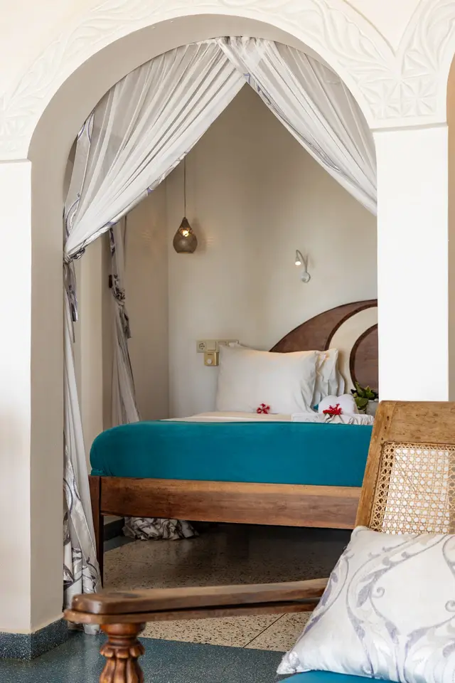 A bed with a turquoise runner under a mosquito net in an arched alcove, with a wooden chair at the edge of the frame