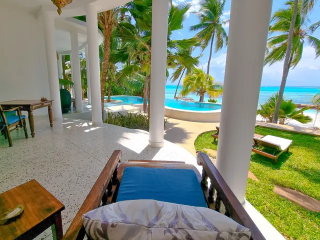A wooden daybed with a blue cushion on a covered terrace, with the pool, palm trees and the ocean beyond