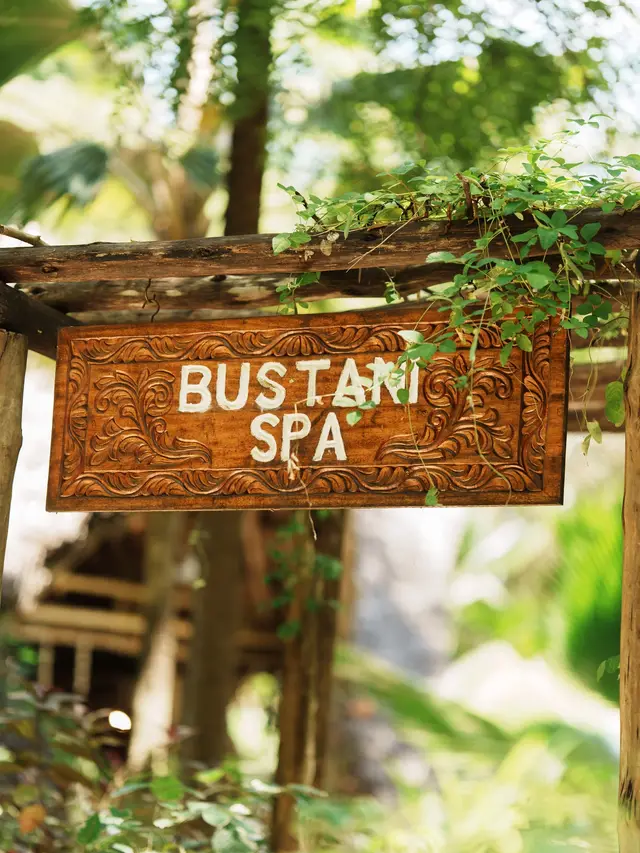 A carved wooden sign reading Bustani Spa, hung among leaves in the garden